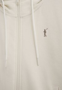Sweat à capuche beige zippé avec cordons blancs, présentant un petit logo brodé d’un joueur de polo sur la poitrine.