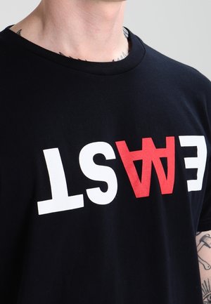 Mand iført en sort T-shirt med ordet "EAST" trykt på hovedet i hvide og røde bogstaver på tværs af brystet.