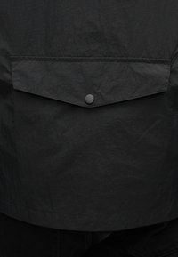 Juoda lengva audinio medžiaga, turinti tekstūruotą paviršių. Gale yra kišenė su atvartėliu ir spaudžiamu užsegimu, išlaikanti paprastą dizainą.