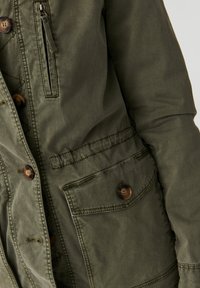 Chaqueta verde oliva hecha de tejido texturizado, con grandes botones, un bolsillo frontal con cremallera y múltiples bolsillos delanteros con detalles de costura.