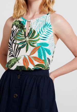 Femme portant un haut sans manches imprimé feuilles tropicales avec un décolleté en forme de goutte et une jupe bleu marine taille haute à boutons, debout avec une main sur la hanche.