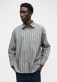 Νεαρός άνδρας με τα χέρια στις τσέπες, φορώντας ένα γκρι και λευκό πουκάμισο με κάθετες ρίγες, μπροστά σε απλό λευκό φόντο.