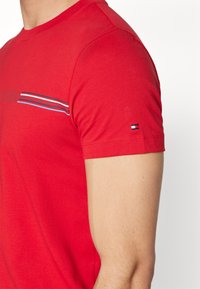 T-shirt in cotone rosso con collo a girocollo, design a righe orizzontali blu, bianche e rosse, e un piccolo logo sulla manica sinistra.
