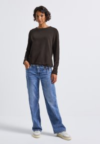 Femme aux cheveux courts et bouclés, vêtue d'un haut à manches longues sombre, d'un jean bleu ample, de baskets blanches, avec une main dans la poche de son jean.