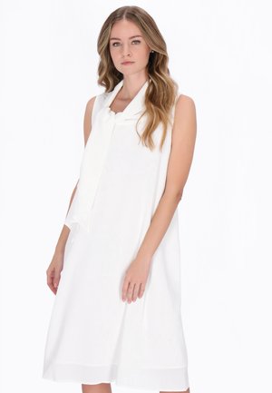 Kobieta o długich falowanych włosach, ubrana w białą, bezrękawnikową sukienkę z kołnierzem i wiązaniem, stoi na tle jednolitego białego tła.