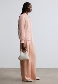 Lyserød bluse med lange ærmer og manchetter med knapper, kombineret med vidde bukser i matchende farve; holder en lille hvid håndtaske.