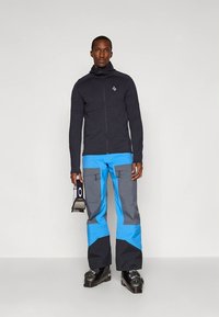 Must zip-up jope kõrge kraega, millele on lisatud sinised ja hallid suusapüksid mitme taskuga. Modell hoiab suusavarustust.