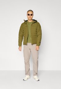 Olivengrønn boblejakke med glidelås foran, sammen med en matchende grøn t-skjorte og lysebeige bukser. Båret med solbriller og joggesko.