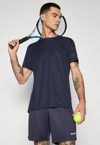 Marinblå sportströja med korta ärmar och subtil logodetalj, matchande shorts, håller en grön tennisboll och racket.