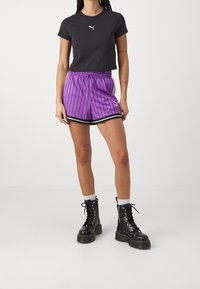 Černé krátké tričko s bílým logem, doplněné fialovými šortkami s proužky, stahovací šňůrkou a černými detaily, nošené s černými kotníkovými botami na tkaničky.
