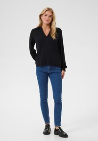 Svart långärmad tröja med V-hals, i kombination med blå smalpassade jeans och svarta loafers, framför en enkel vit bakgrund.