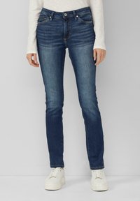 Jeans en denim indigo foncé avec une coupe slim, comportant cinq poches, une fermeture à bouton et des détails de couture subtils, associés à des baskets blanches.
