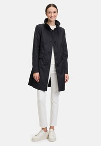Marineblaue, wasserdichte Jacke mit hohem Kragen, Knopfschluss, zwei Fronttaschen und taillierter Passform, kombiniert mit weißen Hosen und Sneakers.