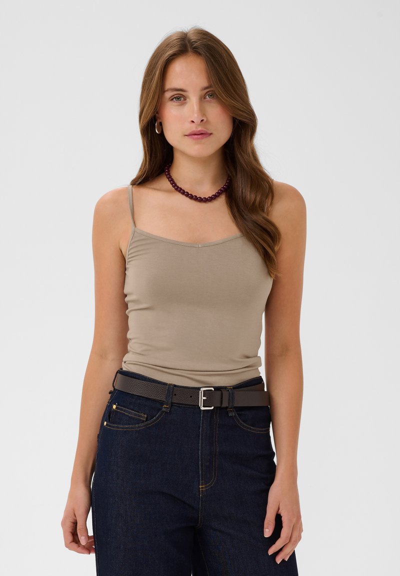 Femme aux longs cheveux bruns portant un débardeur beige, un jean foncé avec une ceinture, un collier de perles bordeaux et des créoles dorées, debout face à l'avant.
