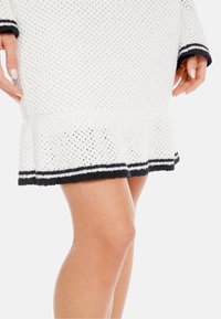 Weißes gestricktes Kleid mit einem strukturierten Muster, das einen ausgestellten Saum sowie schwarz-weiße gestreifte Akzente an den Bündchen und dem unteren Saum aufweist.
