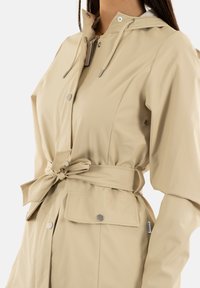 Impermeabile beige con cappuccio regolabile, vita con cintura, bottoni a pressione e tasche laterali; realizzato in materiale liscio e resistente.