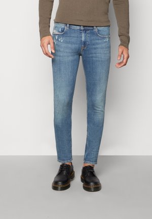 Homme portant un jean bleu slim avec une légère usure subtile et des chaussures noires à lacets, debout devant un fond uni.