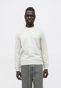 Giovane con capelli intrecciati che indossa una felpa BOSS color avorio e jeans grigi larghi, in piedi davanti a uno sfondo bianco neutro.