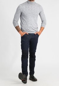 Man med grå polotröja, mörkblå byxor med knälappar, svarta finskor, stående med händerna i fickorna.