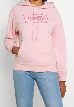 Personne portant un sweat à capuche Levi's rose clair avec poche avant et jean bleu, main dans la poche, sur fond blanc uni.