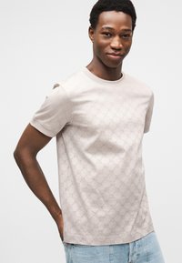 Ανοιχτό μπεζ μπλουζάκι με διακριτό υφασμάτινο μοτίβο, στρογγυλή λαιμόκοψη και κοντά μανίκια, σε συνδυασμό με ανοιχτό μπλε τζιν παντελόνι.