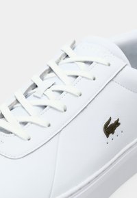 Gros plan d'une basket blanche avec des lacets blancs et un petit logo crocodile foncé sur le côté, sur un fond uni.