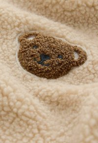 Tissu en polaire beige avec une broderie représentant un visage d'ours brun, comprenant des détails texturés tels qu'une petite bouche et deux oreilles.