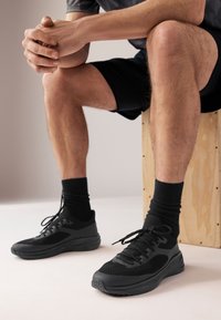 Black athletic shoes with textured mesh and smooth rubber accents, featuring laces and a cushioned sole. Model wearing black socks and shorts.