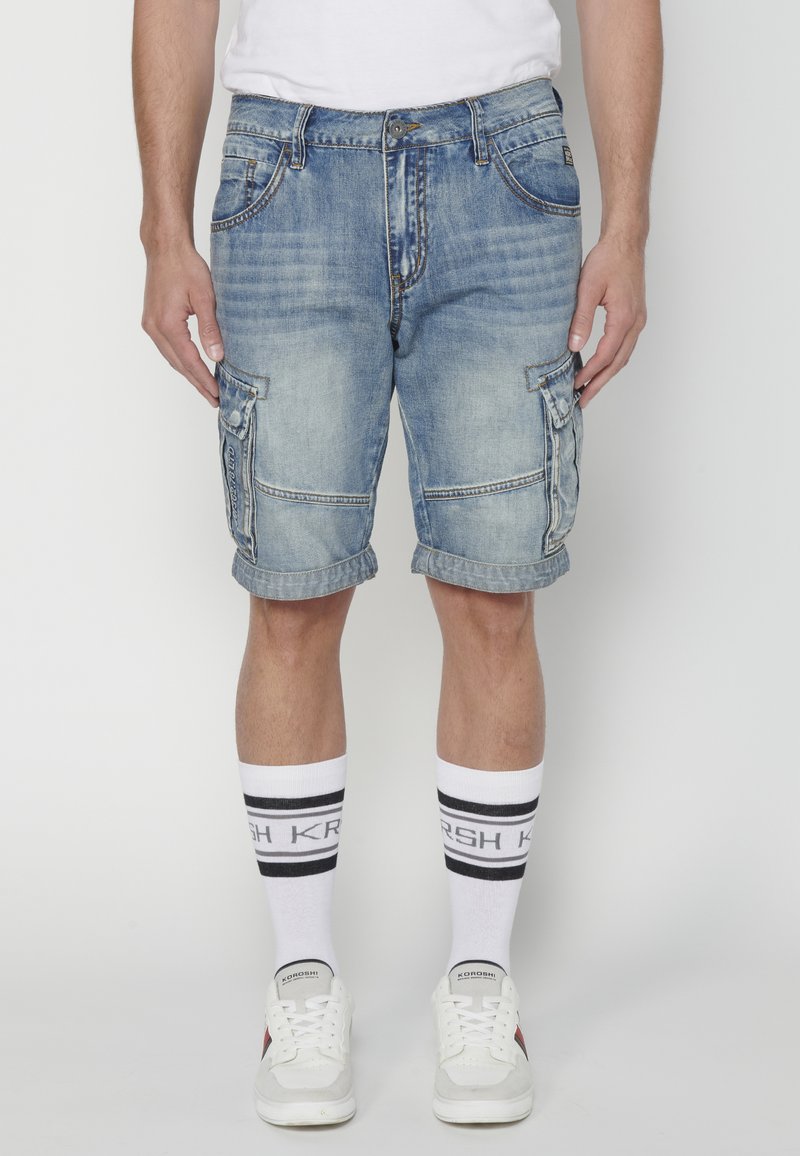 Valkobla denim cargo shorts med flera fickor, rullad fåll och synliga sömdetaljer. I kombination med vita strumpor med svarta ränder.