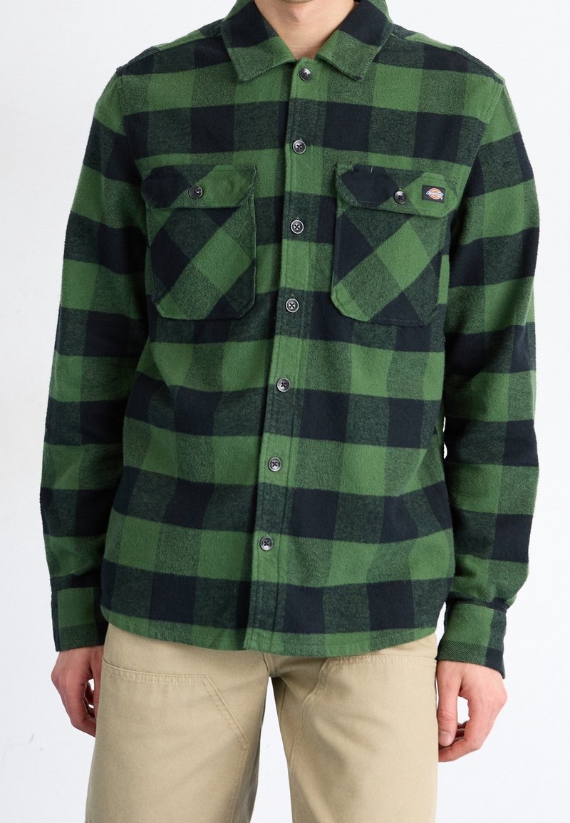 Homme portant une chemise à carreaux verte et noire à boutons avec deux poches poitrine boutonnées et un pantalon beige, montré du cou aux hanches.