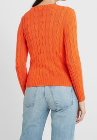 Kobieta w jasnopomarańczowym swetrze w warkocze i jasnoniebieskich dżinsach, przedstawiona od tyłu na jednolitym tle.