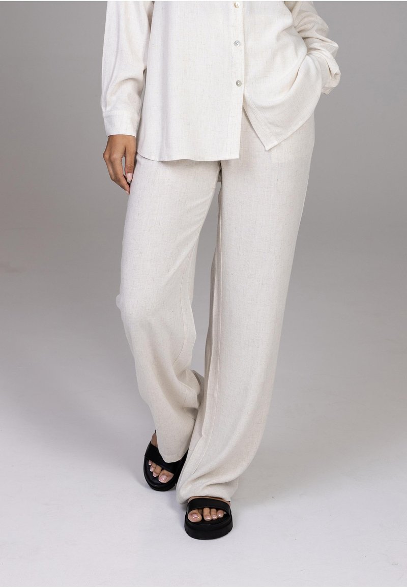 Personne portant un pantalon large beige et une chemise boutonnée à manches longues assortie avec des sandales noires, debout devant un fond neutre.