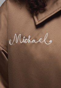 Brązowa satynowa kurtka z dużym białym haftem o kursywie z imieniem "Michael", charakteryzująca się subtelnym połyskiem i gładką fakturą.