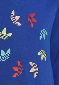 Modrý látkový materiál s několika malými logy Adidas Trefoil v žluté, červené, broskvové a světle modré barvě uspořádanými v rozptýleném vzoru.