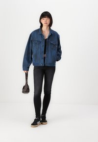 Μπλε τζάκετ από τζιν με δύο μπροστινές τσέπες, φοριέται πάνω από μαύρο τοπ, συνδυασμένο με μαύρα στενά τζιν και μαύρα αθλητικά παπούτσια, κρατώντας μια μικρή καφέ τσάντα.