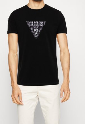 T-shirt en coton noir arborant un graphisme triangulaire avec le texte « GUESS » et un point d'interrogation, sur un fond texturé et à motifs.