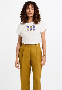 Witte t-shirt met een kleurrijk abstract ontwerp in blauw, goud en bruin, gecombineerd met hoge, mosterdgele broeken.