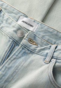 Világoskék farmernadrág cipzáros záródással, ezüst színű fémalkatrészekkel és egy fehér címkével, amelyen "JACK&JONES" felirat található. A gomb körüli rész kopott hatású.