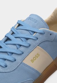 Αθλητικό παπούτσι από γαλάζιο σουέντ με κορδόνια στο ίδιο χρώμα, μπεζ πλαϊνή επένδυση, χρυσό λογότυπο "BOSS" και σόλα χρώματος καραμέλας, σε διαγώνια όψη.
