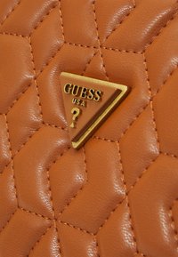 Borsa a mano in pelle trapuntata marrone con un'emblema dorato triangolare con la scritta "GUESS USA" e un punto interrogativo. Cuciture dettagliate visibili.