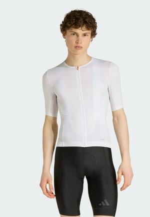 Giovane uomo con capelli ricci che indossa una maglia da ciclismo bianca aderente e pantaloncini imbottiti neri su uno sfondo semplice.