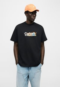 Άνδρας που φοράει πορτοκαλί καπέλο, μαύρο μπλουζάκι με στάμπα και ανοιχτόχρωμα τζιν, στέκεται με τα χέρια στις τσέπες, κοιτάζοντας προς τα πλάγια.