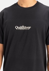 Camiseta de algodón negra con un logo blanco de Quiksilver que incluye un diseño de ola y el texto "The Original Surf Co Since 1969" debajo.