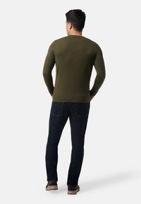 Mann, der mit dem Rücken steht, trägt einen olivgrünen Pullover, dunkelblaue Jeans und braune Schuhe vor einem einfachen hellen Hintergrund.