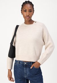 Beige strikkegenser med ribbestrikket kant og mansjetter, med rund hals og knapppynt på skulderen, brukt sammen med høytlivd jeans.