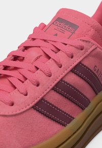Ροζ αθλητικό παπούτσι Adidas Gazelle με καουτσούκ υφή, σκούρες ροζ ρίγες, κορδόνια στο ίδιο χρώμα και σόλα από καουτσούκ.