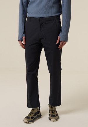 Mann trägt schwarze, gerade geschnittene Hose, blaues, langärmliges, strukturiertes Hemd und grün-schwarze Wanderschuhe, steht vor neutralem Hintergrund.