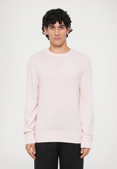 Un jeune homme avec des cheveux bouclés et foncés portant un pull en col rond rose clair et un pantalon noir, se tenant devant un fond blanc uni.