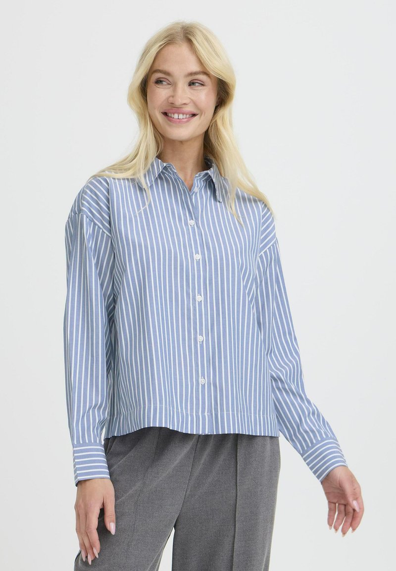 Lächelnde Frau mit langen blonden Haaren, die ein blau-weiß gestreiftes Hemd und graue Hose trägt, vor einem einfarbigen Hintergrund stehend.