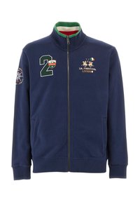 Marinblå zip-jacka i mjukt tyg; har grön och röd krage, broderade logotyper och framfickor. Formen är figurnära med ribbad mudd.